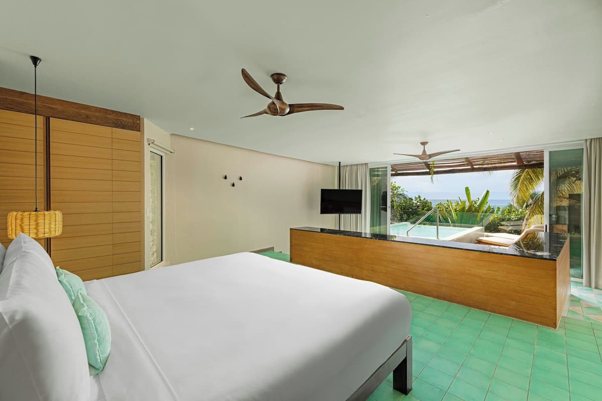 Habitación de hotel con cama, ventiladores de techo, televisor, balcón con piscina y vista al océano.