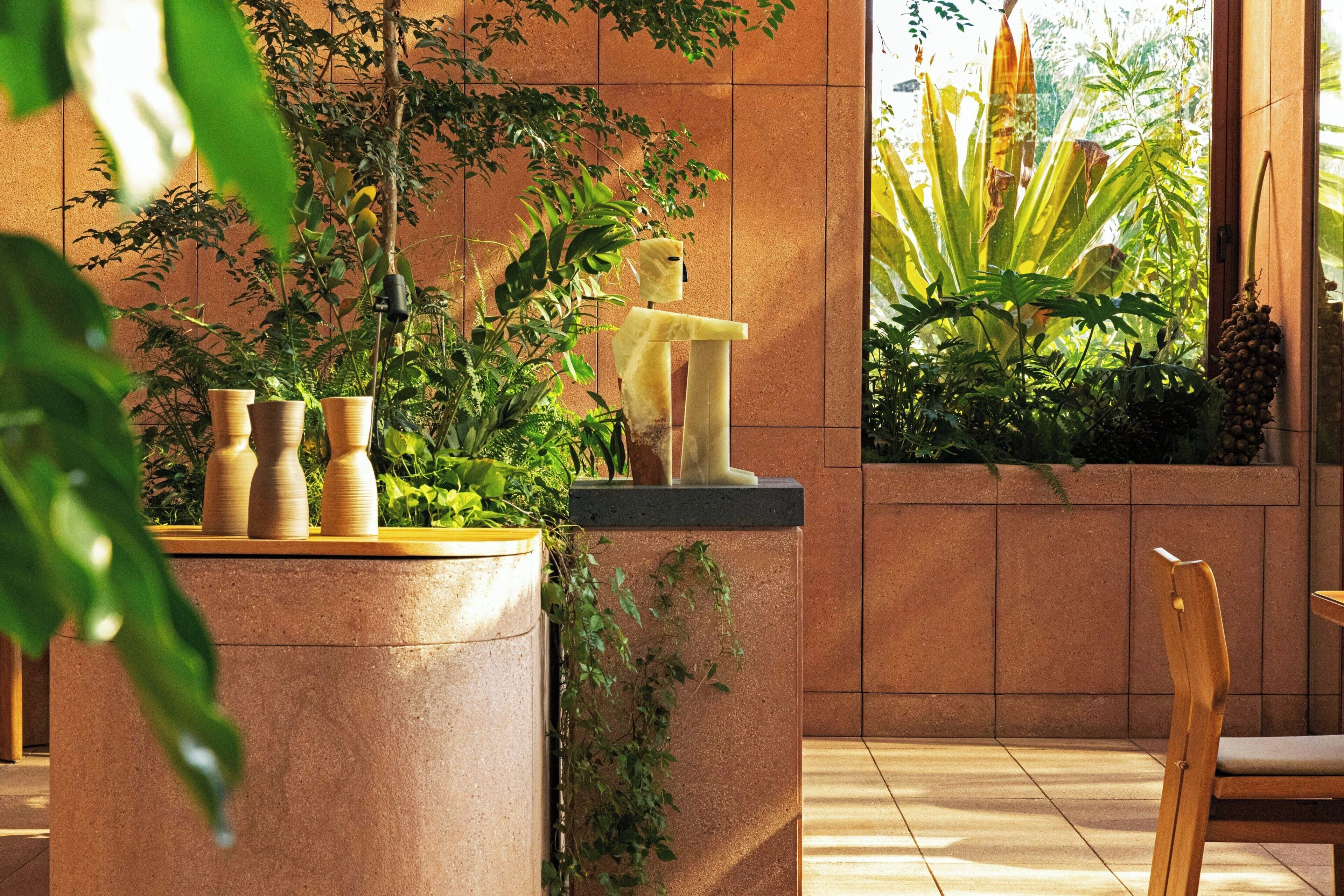 Un espacio interior moderno con una maceta grande llena de plantas diversas, una silla de madera y una escultura en la pared.