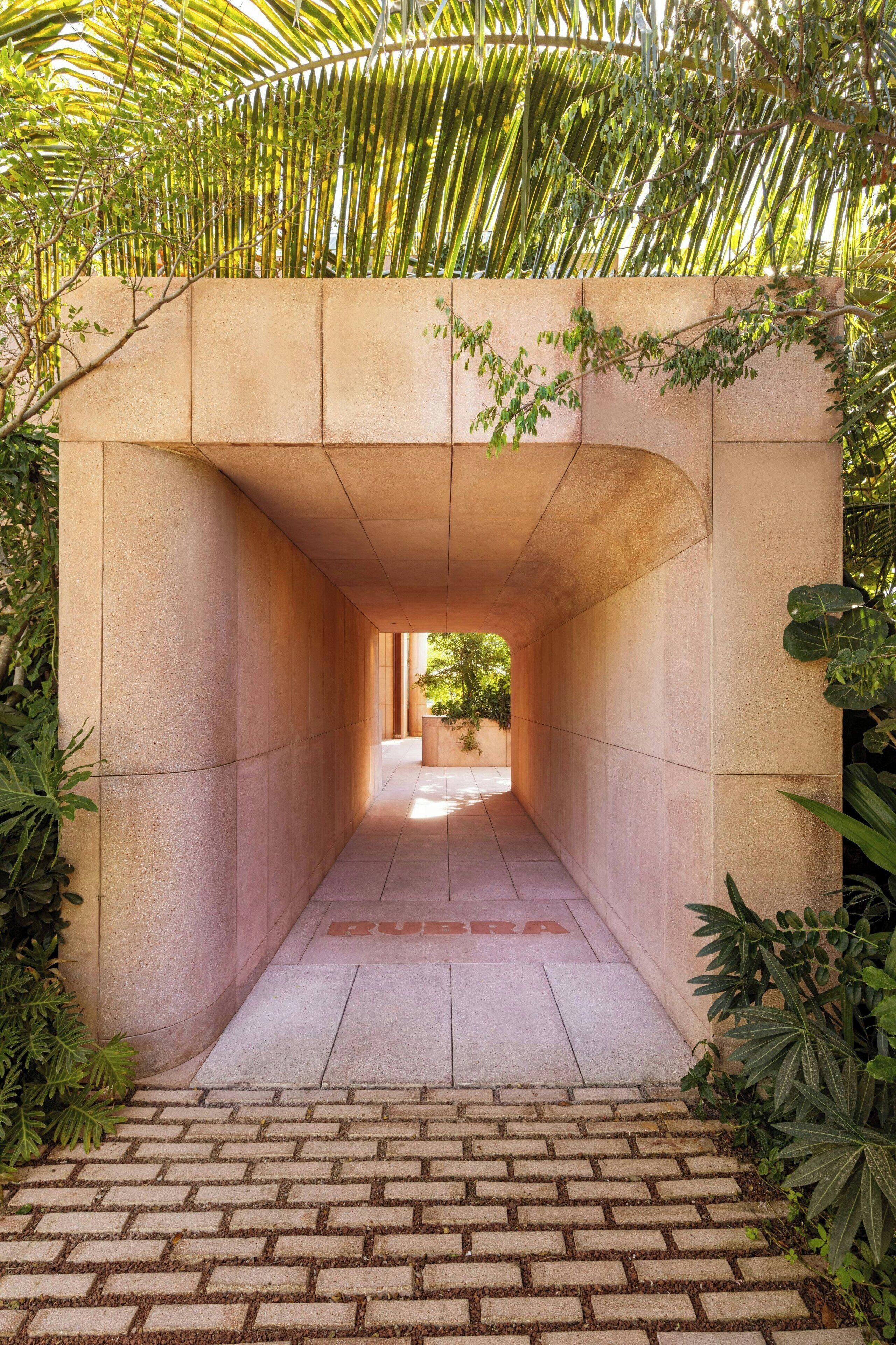A pathway leading through a covered area with a brick floor, surrounded by greenery and a concrete wall.
