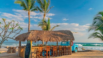 A beach bar with a thatched roof, wooden chairs, and a surfboard.