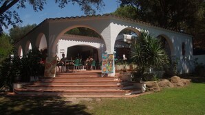 A covered outdoor seating area with tables and chairs, surrounded by greenery and a white building with arches. - Camping Internacional Palamos (Palamós)
