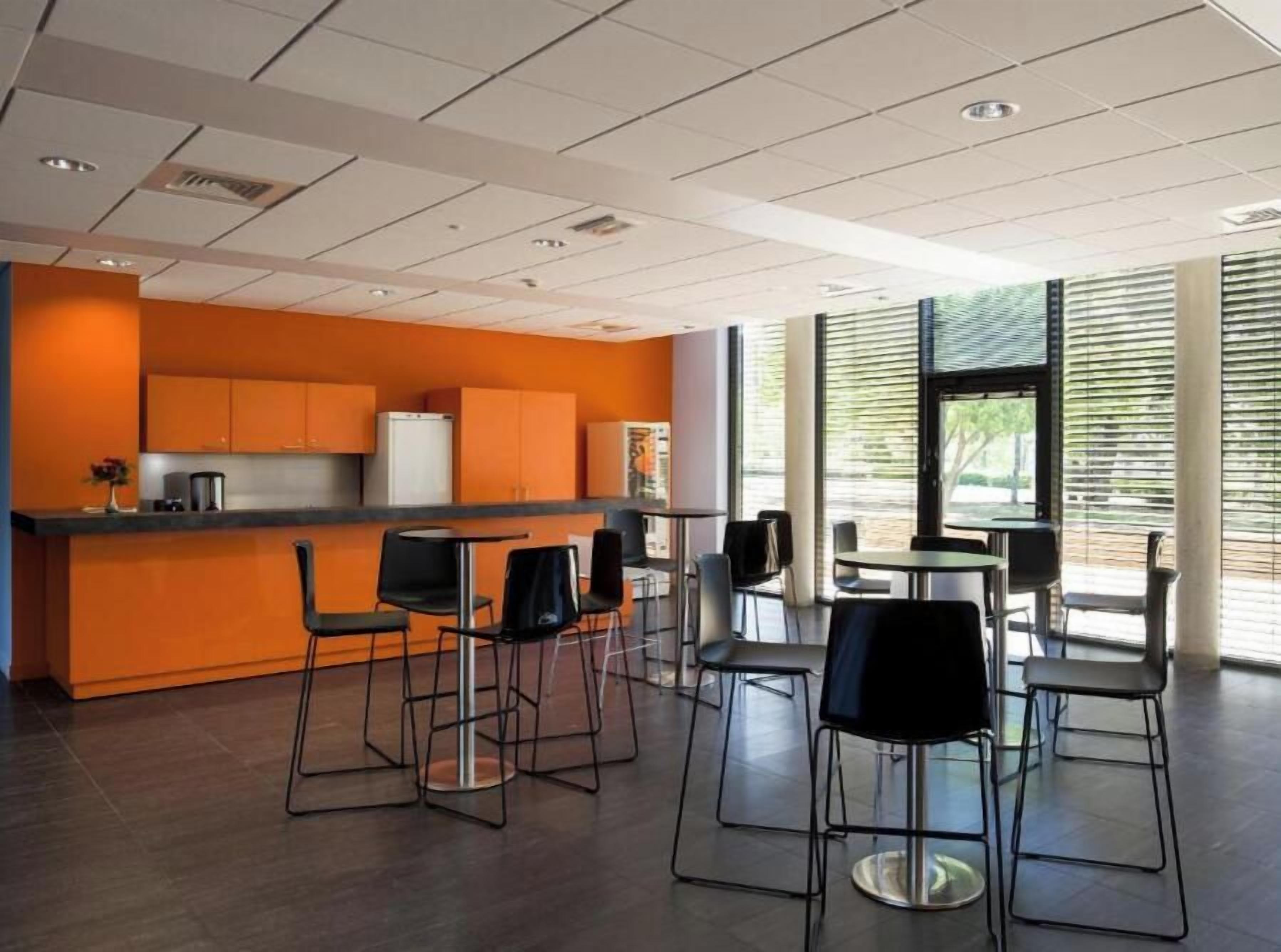 A modern kitchen area with bar stools and a dining table, large windows with blinds, and a ceiling with recessed lighting.
