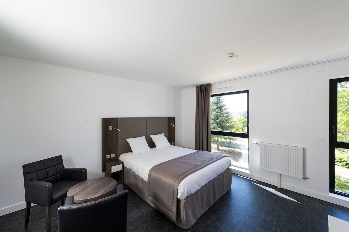 A modern hotel room with a large bed, a desk, a chair, and a window with a view of trees.