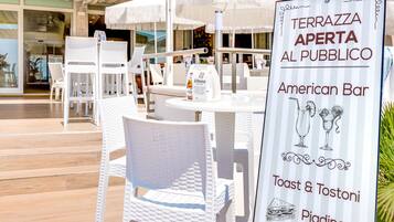 Un'area esterna con sedute e tavoli in vimini bianco, un cartello pubblicitario di un American Bar, Toast & Tostoni, e Piadine.