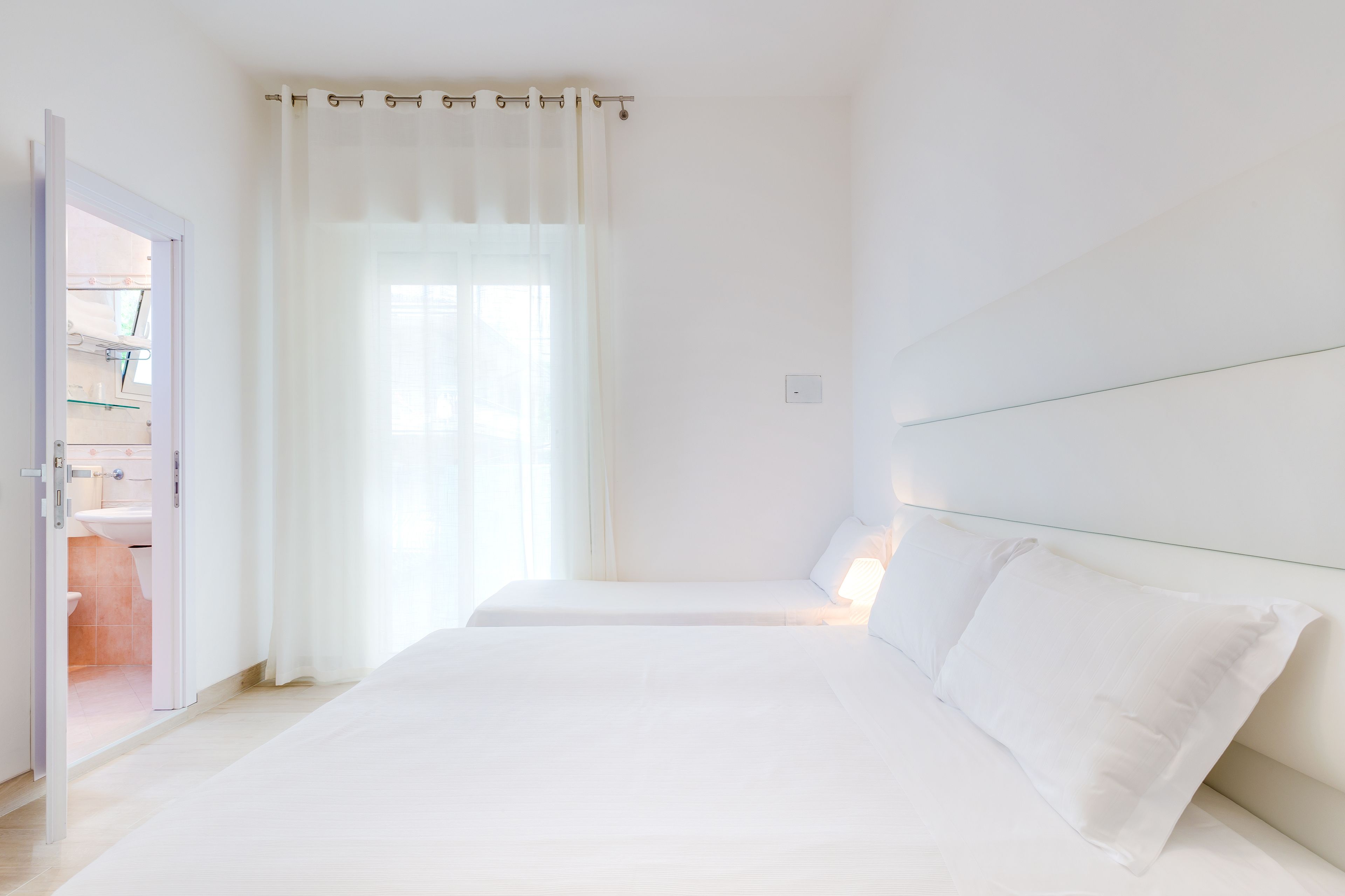A minimalist hotel room with two beds, a bathroom visible through an open door, and a large window with sheer curtains.