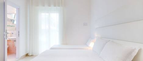 A minimalist hotel room with two beds, a bathroom visible through an open door, and a large window with sheer curtains.