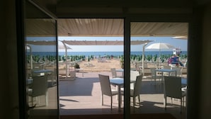 A beach view from a covered patio with white chairs and tables, umbrellas, and a clear sky. - Hotel Bellariva (Jesolo)