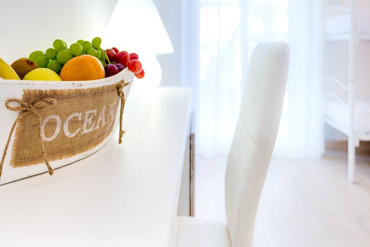 A white room with a table holding a fruit bowl labeled "OCEAN" and a white chair.