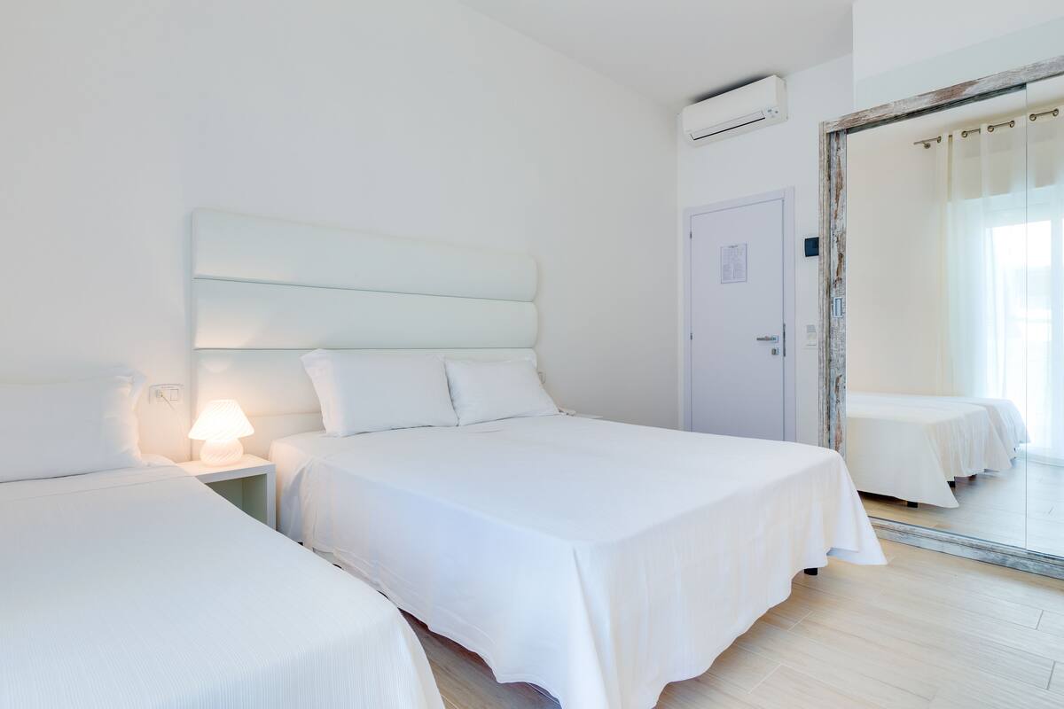 A hotel room with two beds, a nightstand with a lamp, a large mirror, and an air conditioning unit.