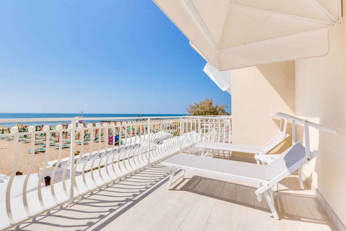 A balcony with white sun loungers, a view of the beach, and a clear blue sky.