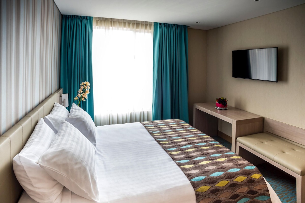 Une chambre d’hôtel avec un lit, un bureau, une banquette, une télévision et une fenêtre avec des rideaux.