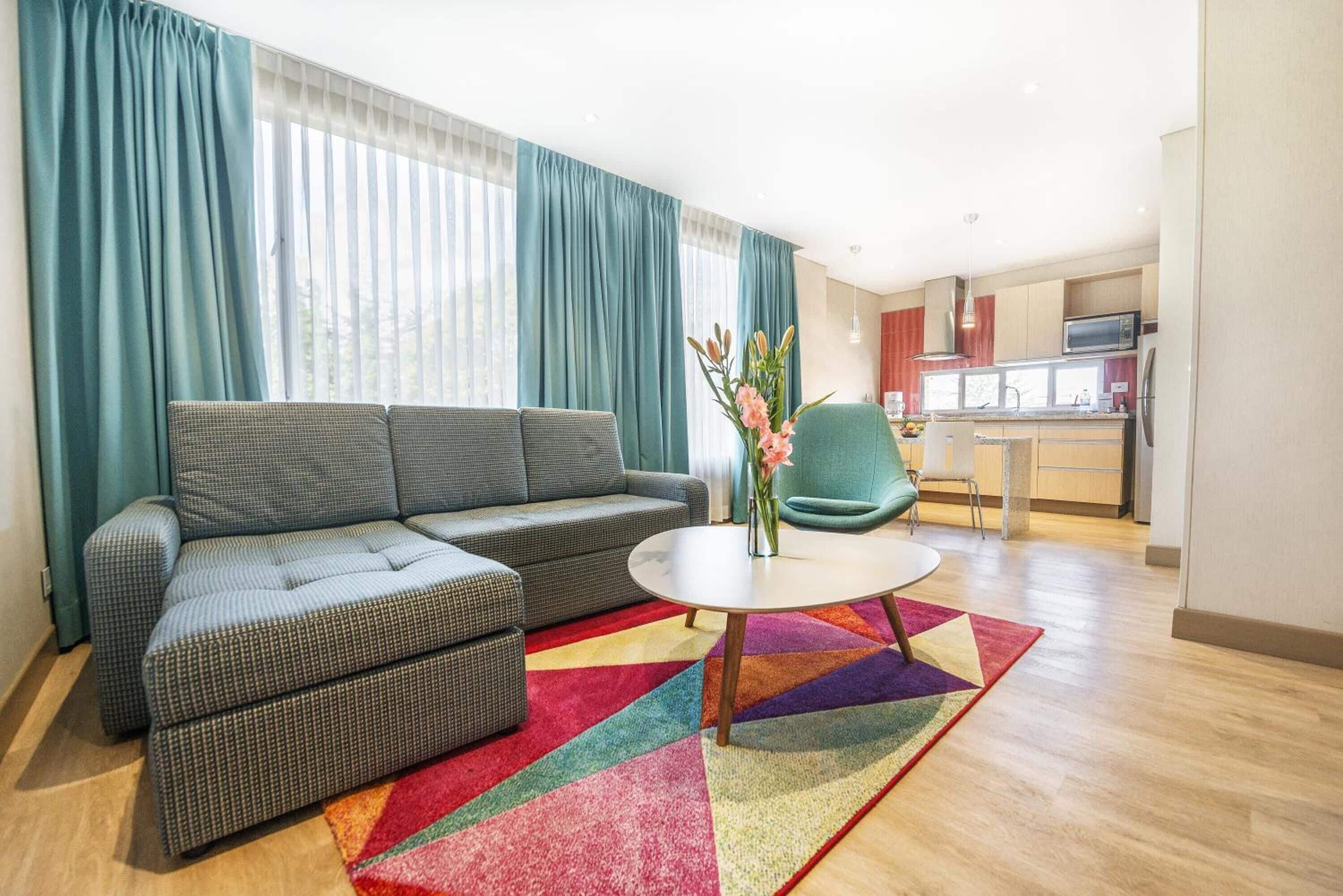 A modern living room with a grey sofa, a round coffee table, and a colorful rug. There is a kitchen area with a microwave and a sink in the background.