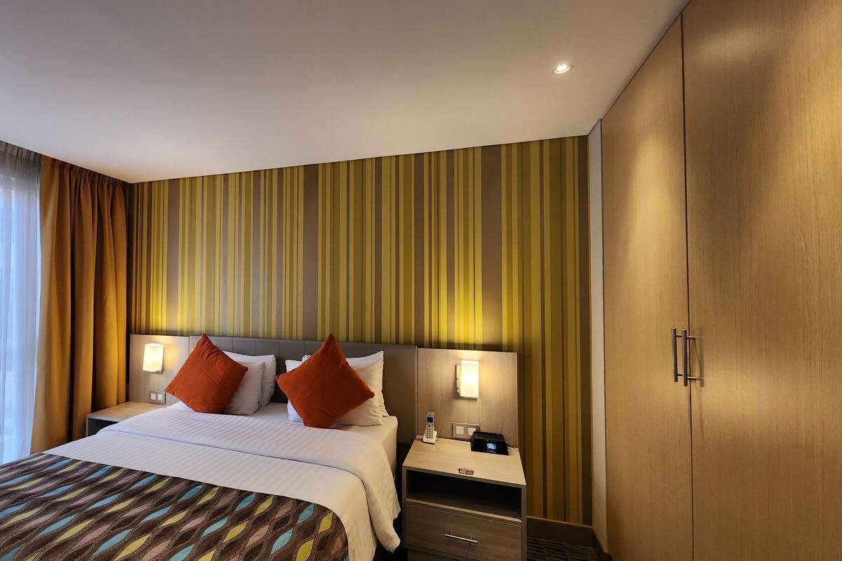 Habitación de hotel con una cama, dos mesitas de noche con lámparas, un armario de madera y una pared a rayas.