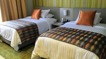 Ein Hotelzimmer mit zwei Betten, jedes mit einem gemusterten Bettbezug und passenden Kissen. Zwischen den Betten befindet sich ein Nachttisch mit einer Lampe und einem Telefon.