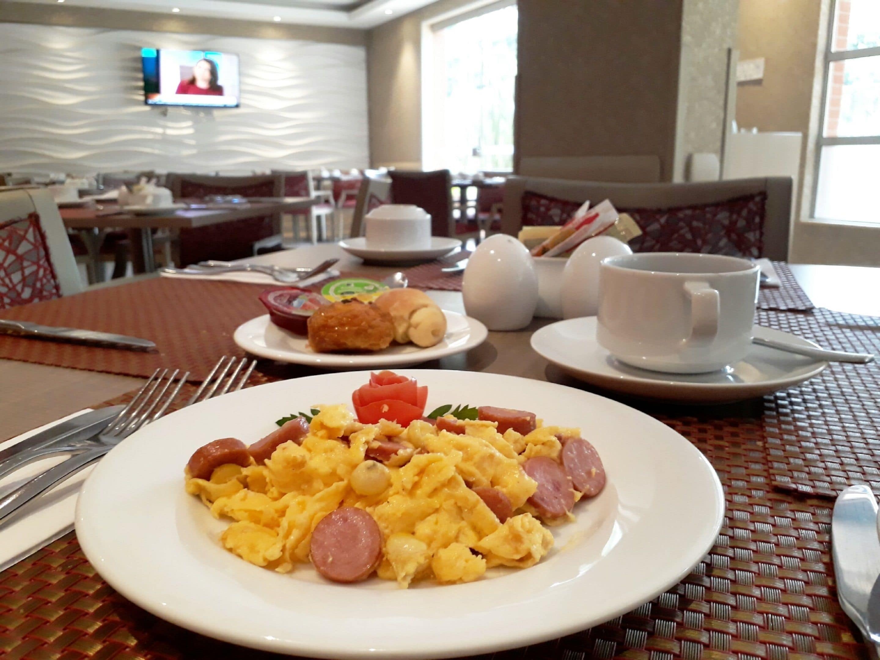 A breakfast setting with scrambled eggs, sausage, and a side of toast. There are dining tables with chairs, a television screen, and a window in the background.