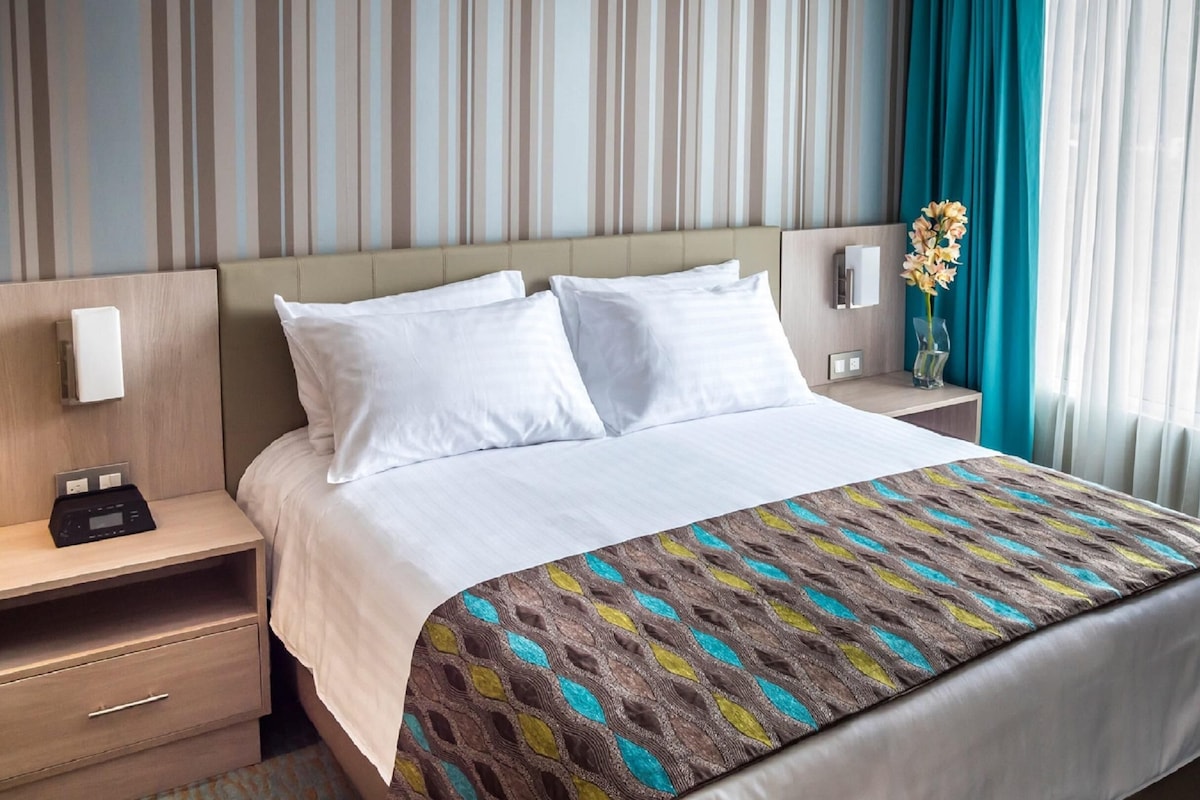 Une chambre d’hôtel comprenant un lit, deux tables de chevet, une petite table, un téléphone fixé au mur et une fenêtre avec des rideaux.