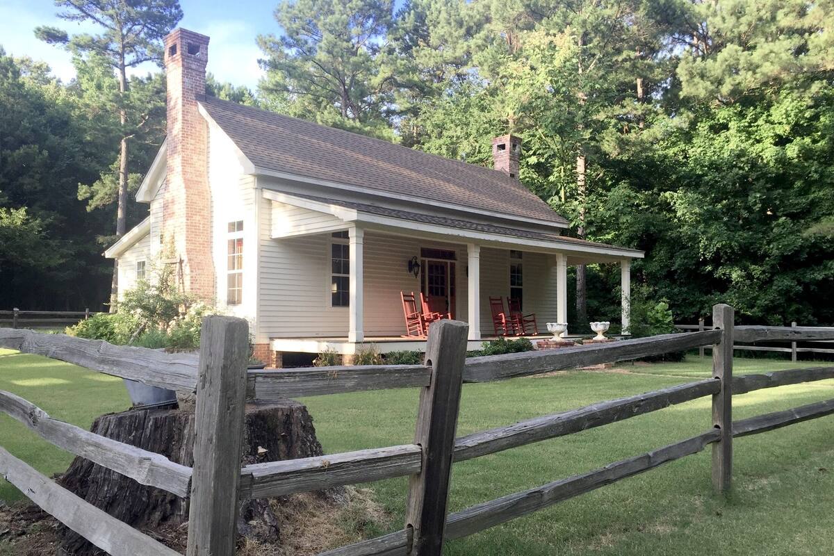 A two-story house with a brick chimney, a porch with red chairs, and a wooden fence.