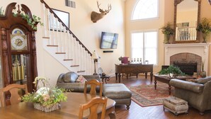 A spacious living room with a wooden staircase, a mounted deer head, a fireplace, and a television on the wall.