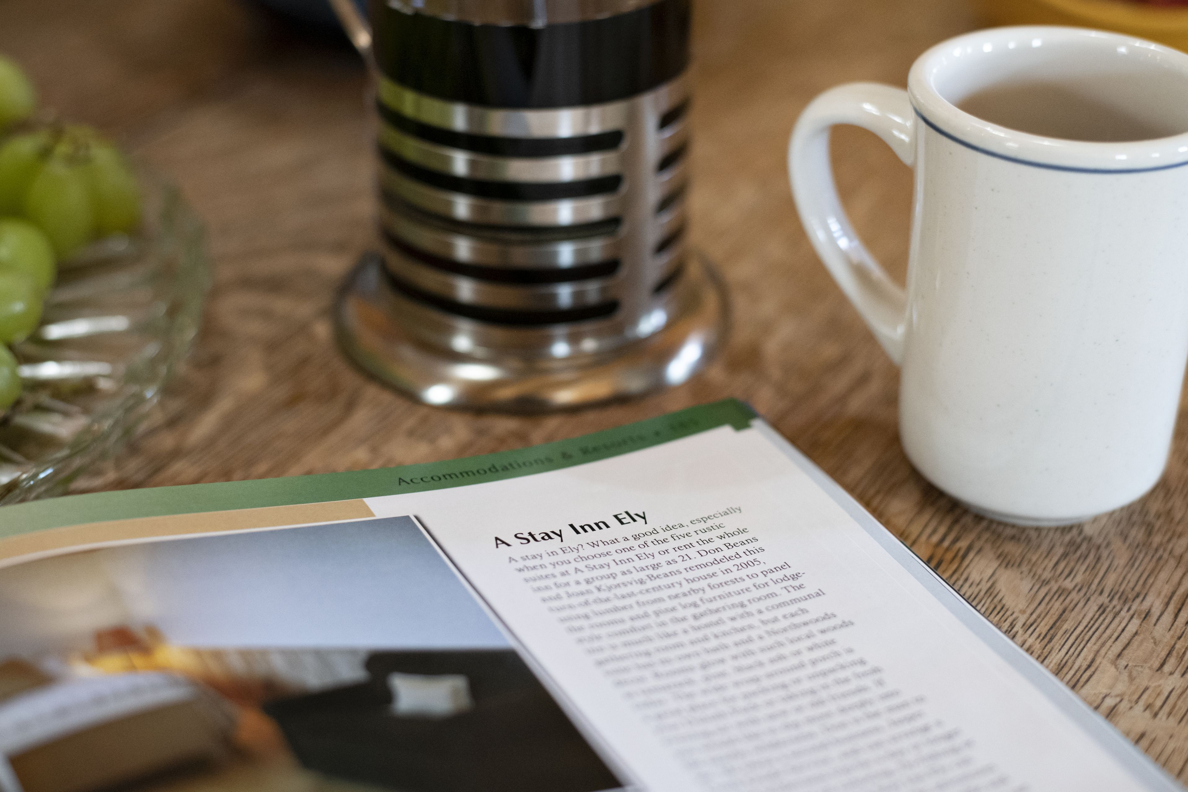 Una taza blanca con borde azul, una jarra de vidrio y un folleto sobre una mesa de madera.