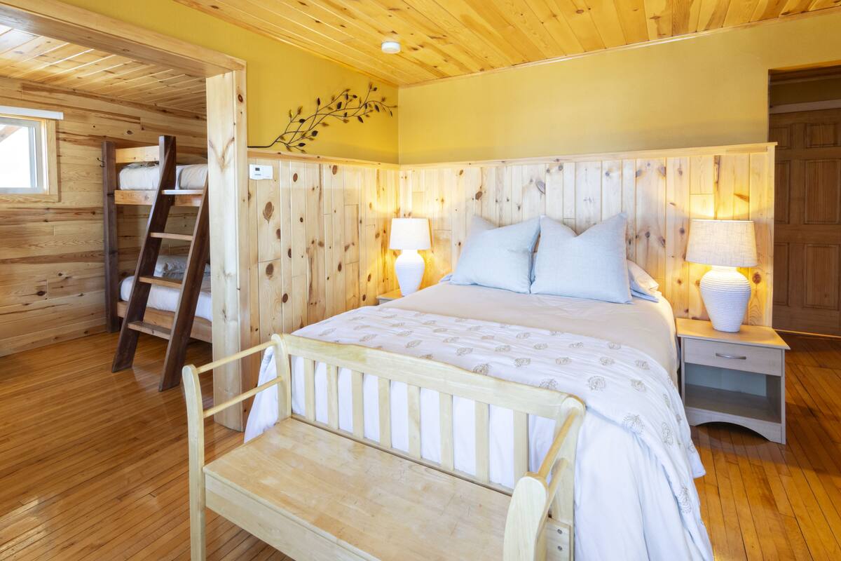 A wooden cabin bedroom with a bunk bed, a ladder, a bedside table with a lamp, and a nightstand.