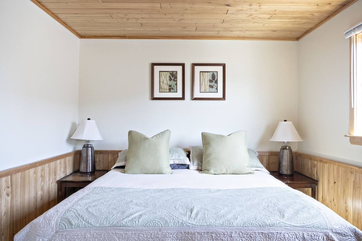 A bedroom with a wooden ceiling, two framed pictures on the wall, a bed with a patterned bedspread, two bedside lamps, and a window on the right.