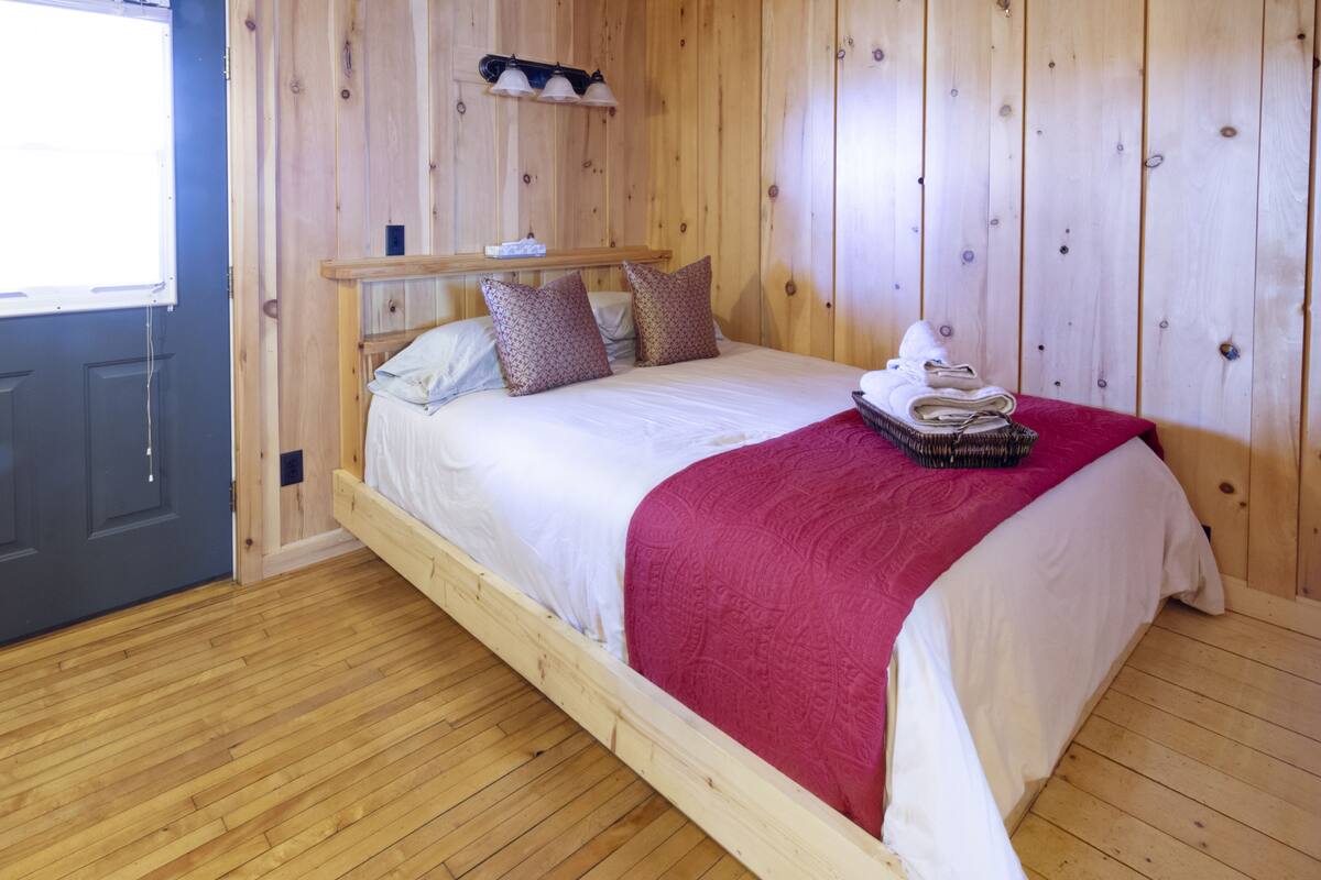 A wooden bed with white and red bedding, a wooden floor, and a wooden wall with a window.