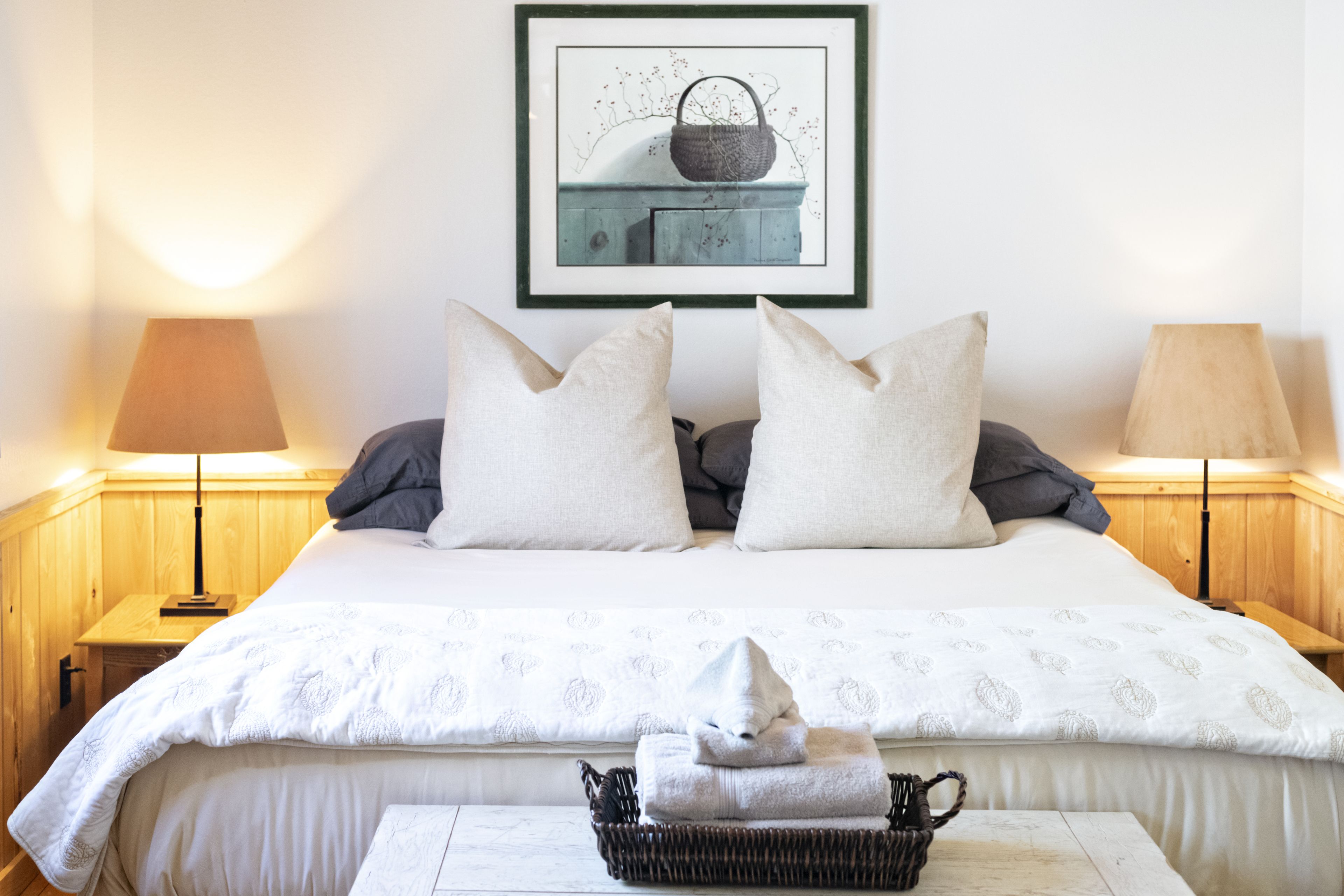 Un lit bien fait, avec une couette blanche, deux oreillers et un panier rempli de serviettes pliées, posé sur une table de chevet en bois.