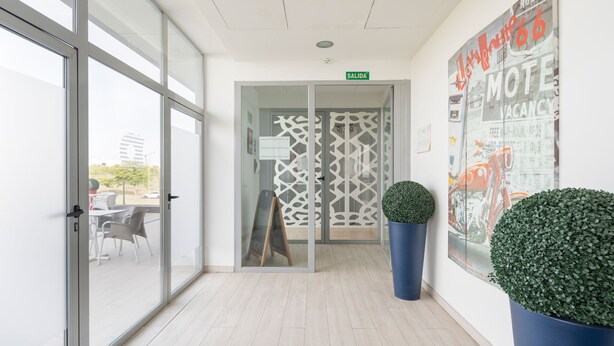 A modern hotel lobby with glass doors leading to a seating area, a large poster on the wall, and two potted plants. - Hotel Route 42 (Illescas)