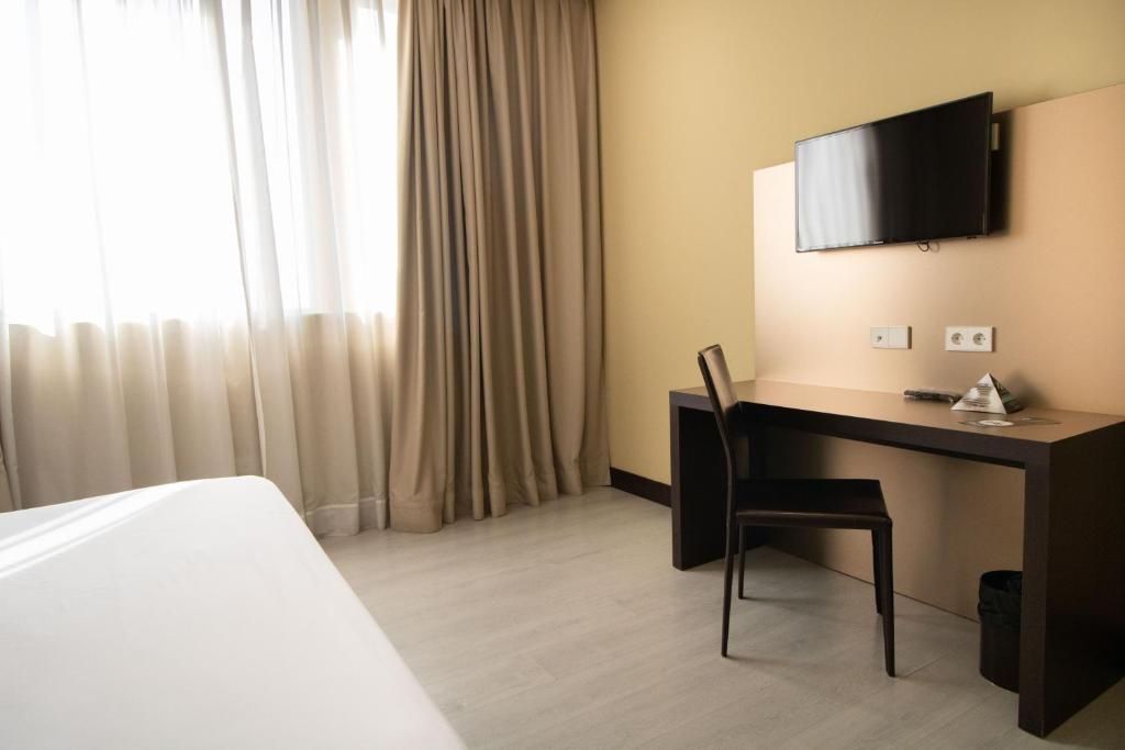 A hotel room with a bed, a desk, a chair, a television mounted on the wall, and a window with curtains.