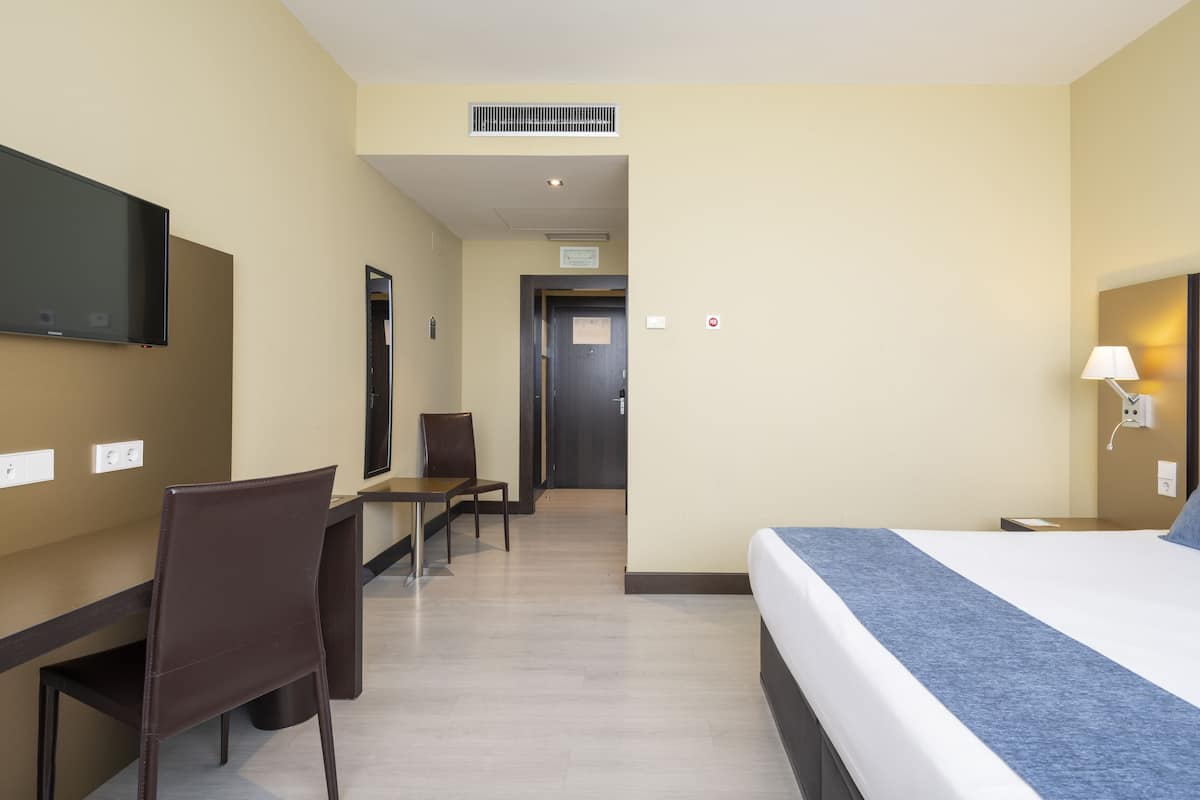 A hotel room with a bed, desk, chairs, a television, and a lamp.