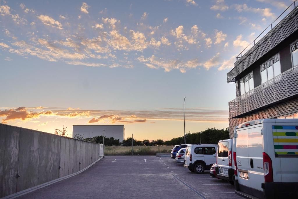 Un estacionamiento con varios vehículos, un muro de concreto y un edificio con fachada metálica.