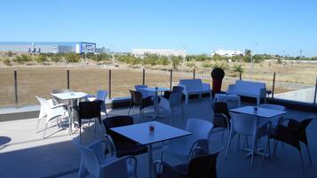 Un patio al aire libre con mesas y sillas, vista al mar y un edificio distante.