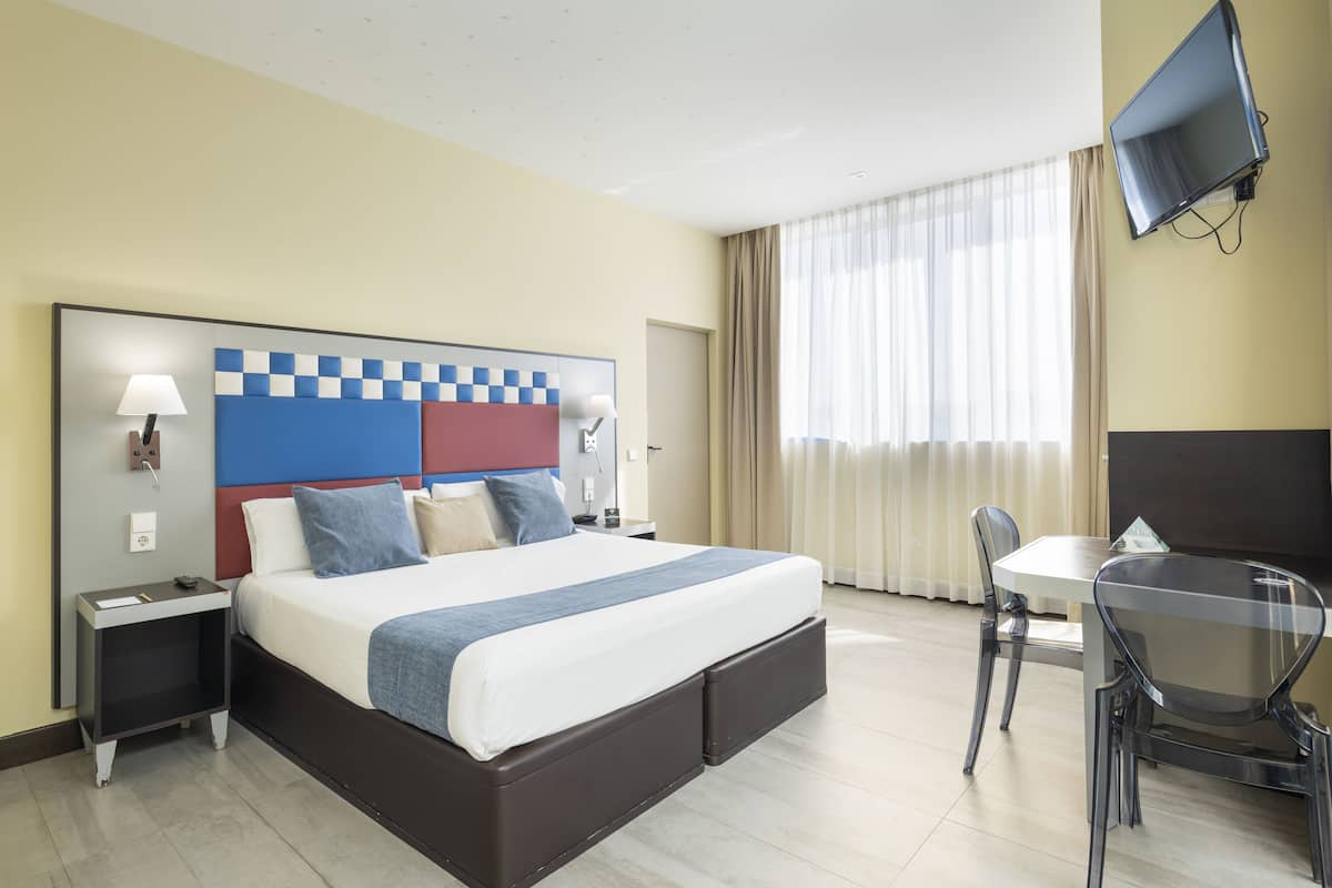 A hotel room with a bed, bedside tables, a TV, a desk, and a chair.