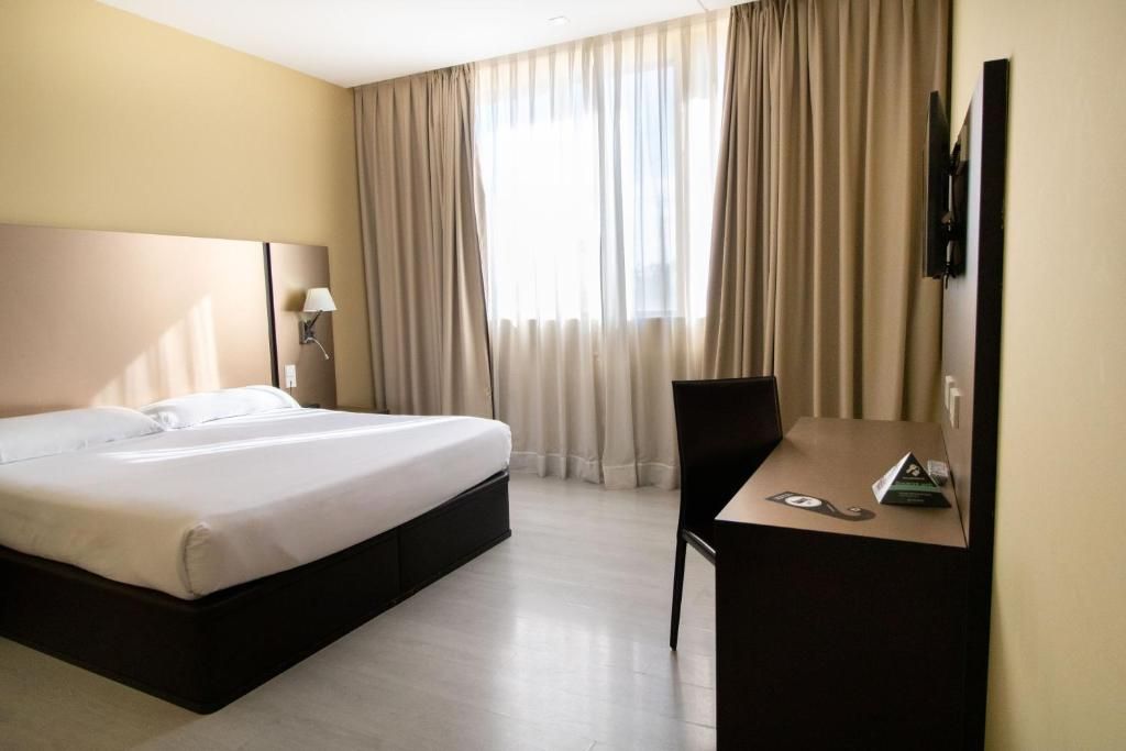 A hotel room with a bed, a desk, a chair, a TV, and a window with curtains.