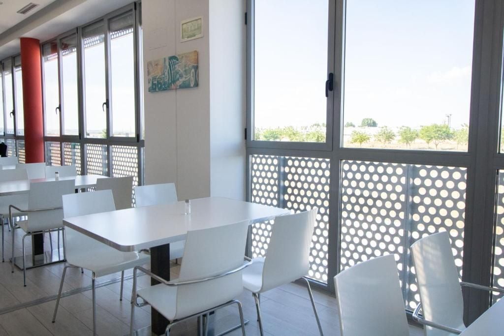 Un espace repas moderne avec des tables et des chaises blanches, de grandes fenêtres et un pilier rouge.