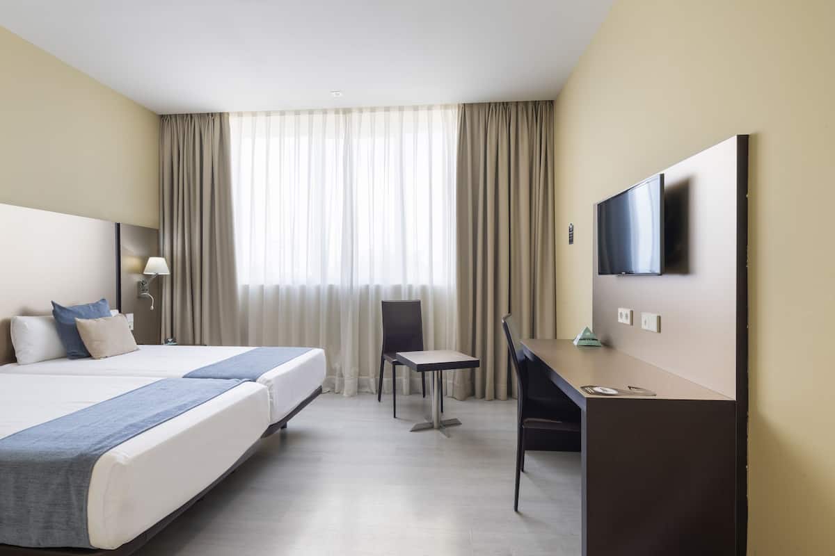 A hotel room with two beds, a desk, a chair, a TV, and a window with curtains.