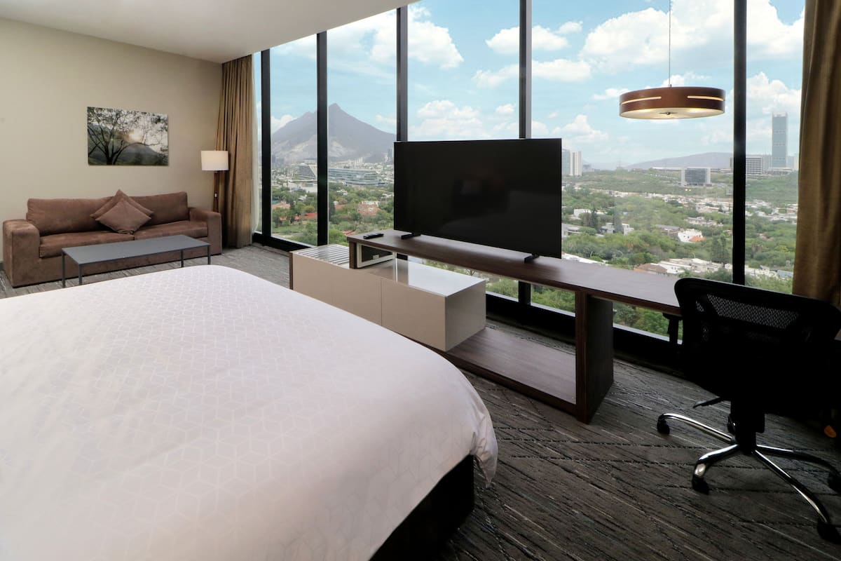 A hotel room with a large bed, a TV on a desk, a sofa, and a view of a cityscape with mountains.