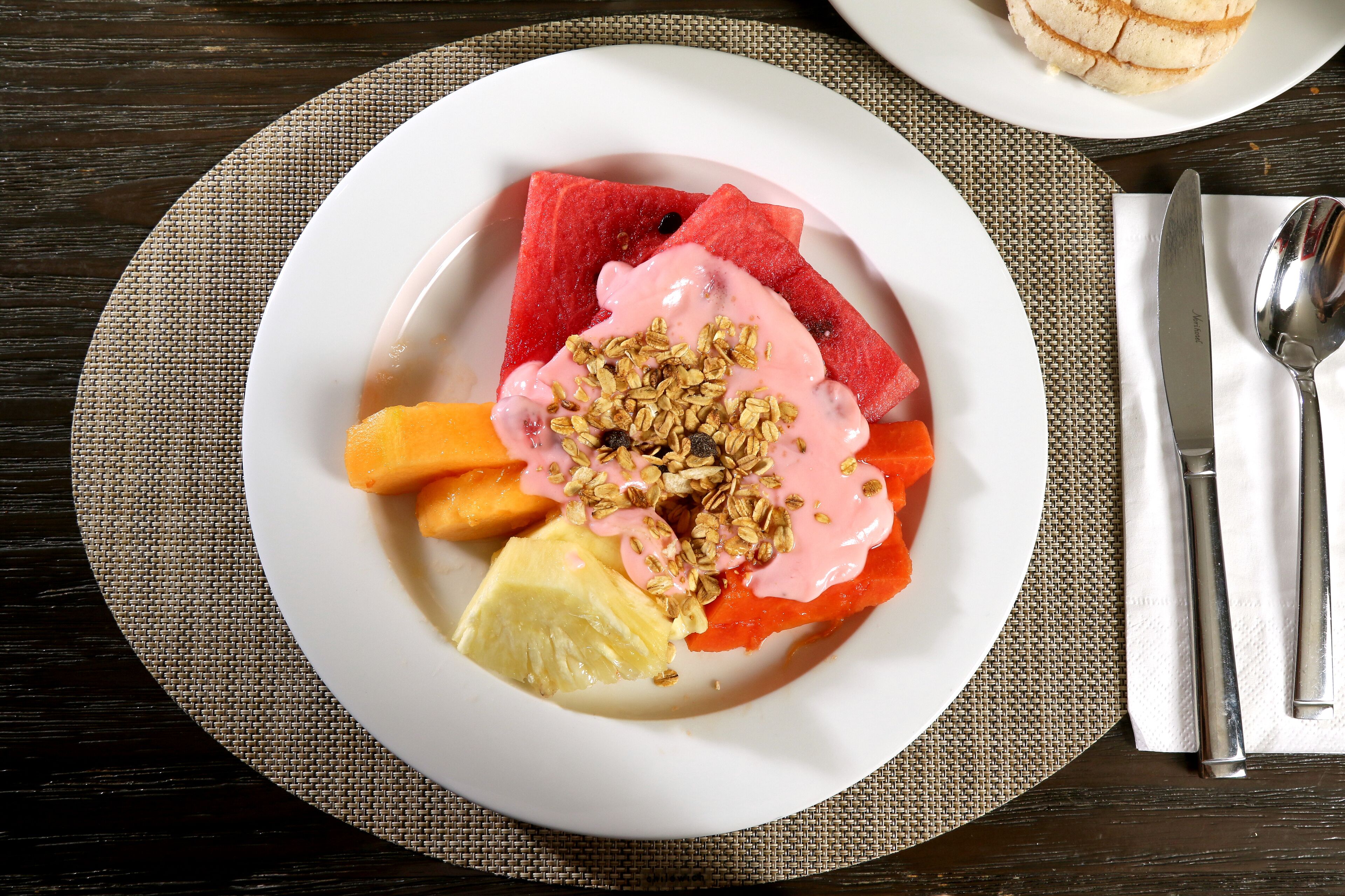 Een fruitsalade met watermeloen, sinaasappel, ananas en granola, geserveerd op een wit bord.