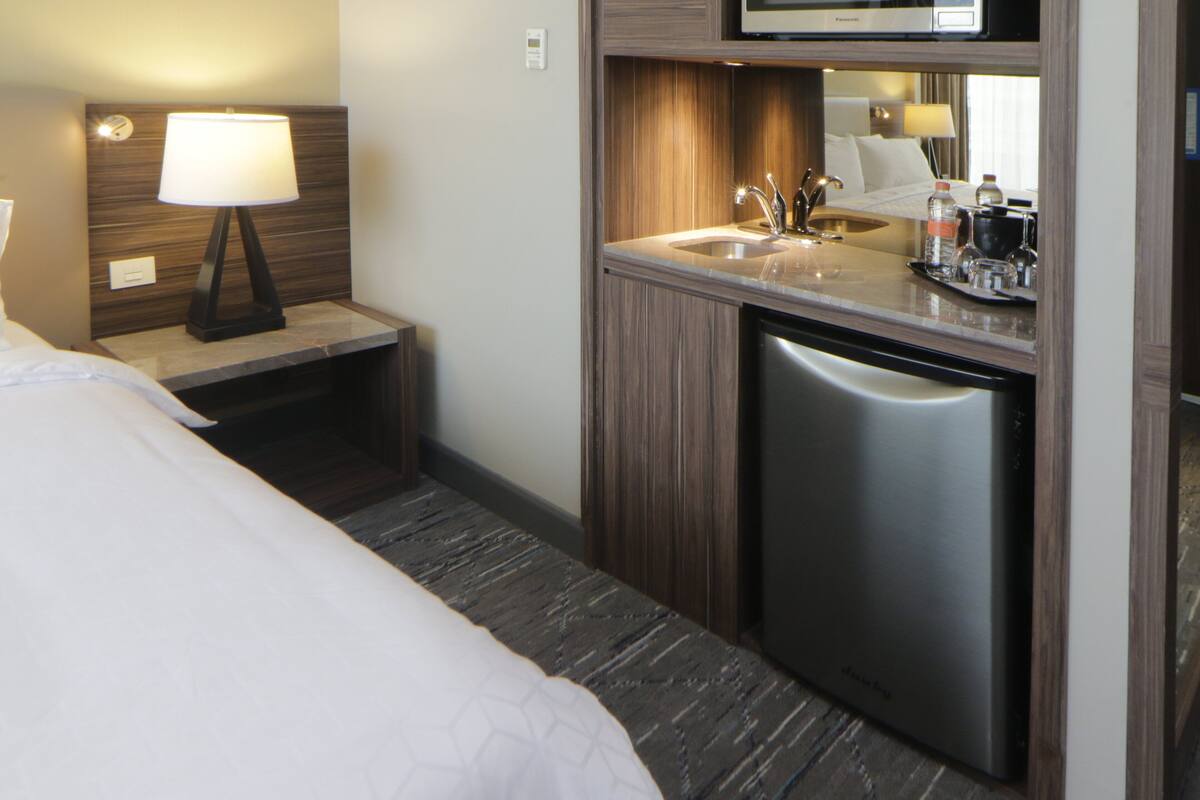 A compact hotel room with a bed, a bedside lamp, a small sink, and a built-in dishwasher.