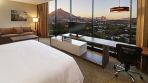 A hotel room with a large bed, a desk with a computer, a TV, and a view of a cityscape and mountain. - Holiday Inn Express & Suites Monterrey Valle by IHG (San Pedro Garza García)