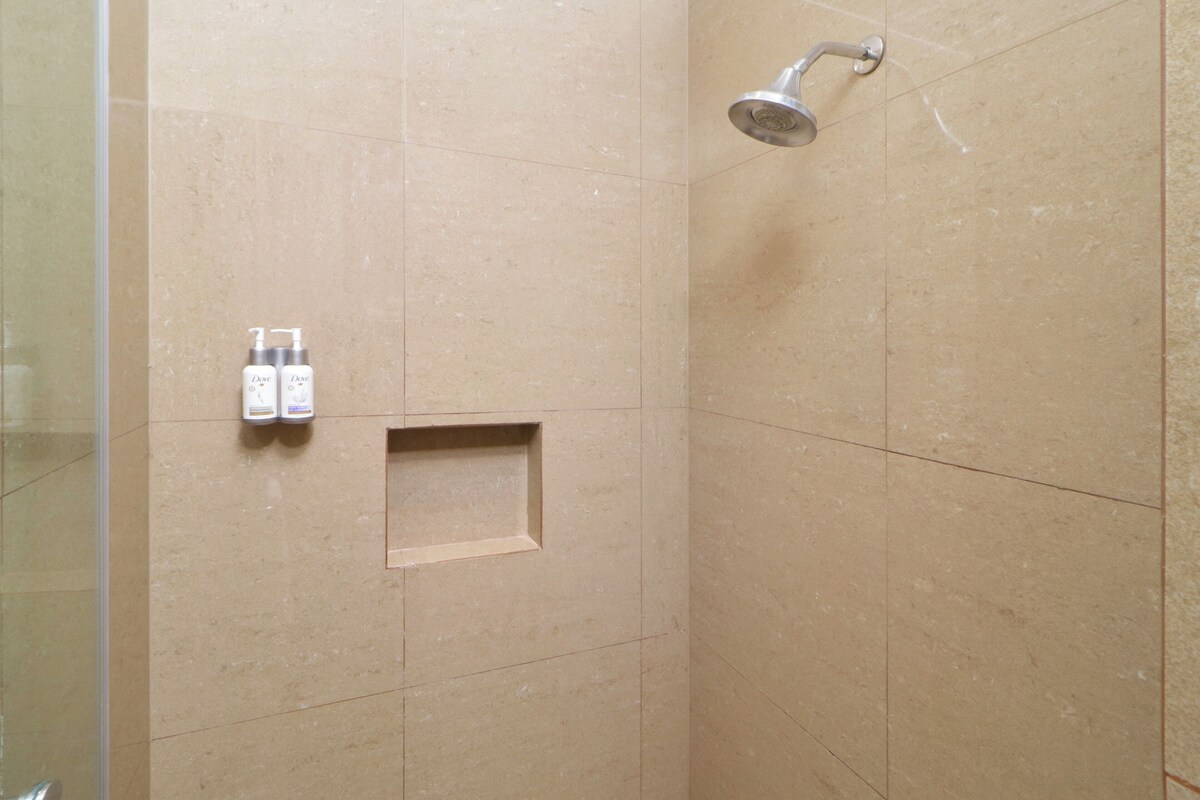 A shower area with beige tiles, a showerhead, and two bottles of Dove body wash on the wall.