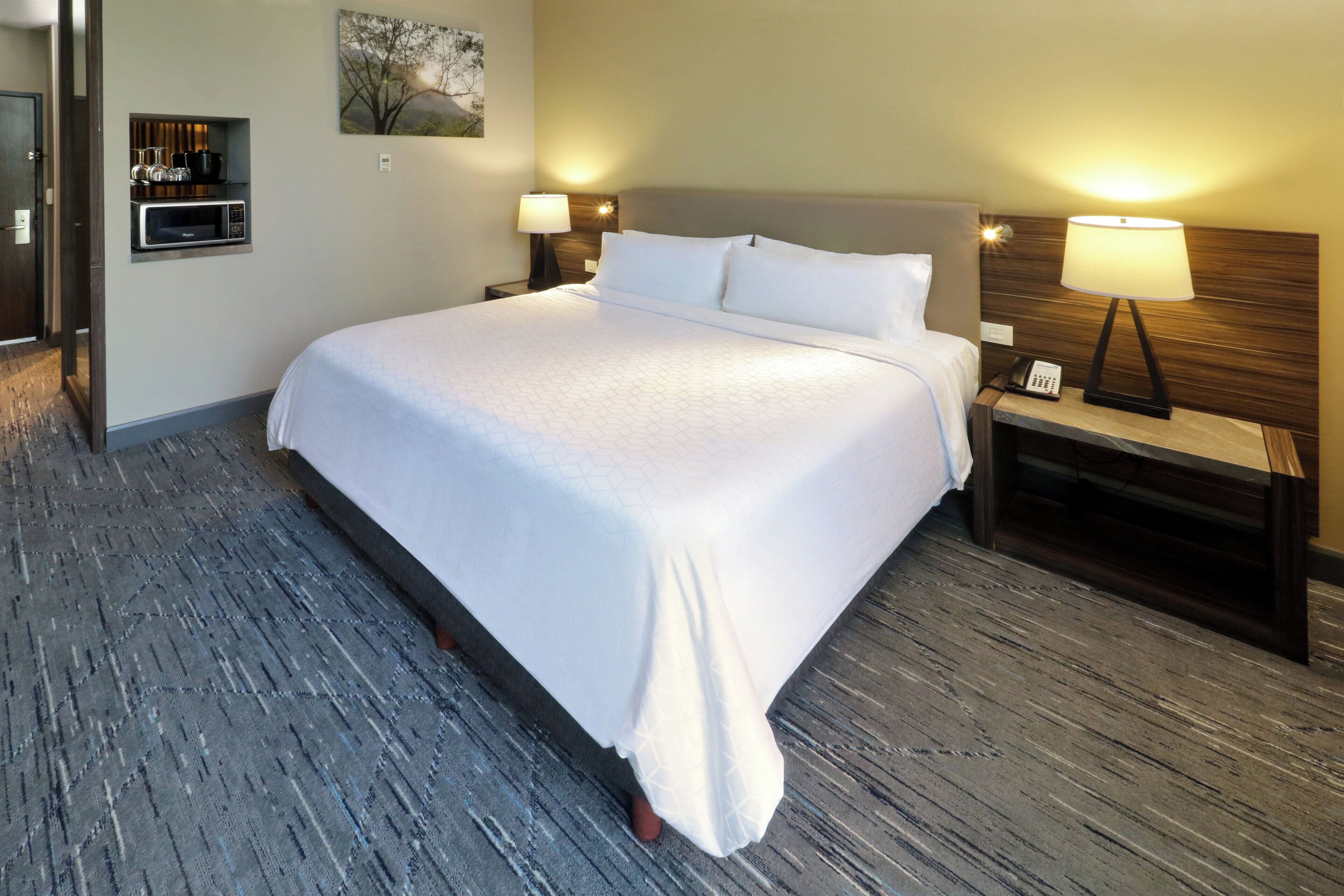 A hotel room with a large bed, two bedside tables with lamps, a wall-mounted telephone, and a small shelf with glassware.