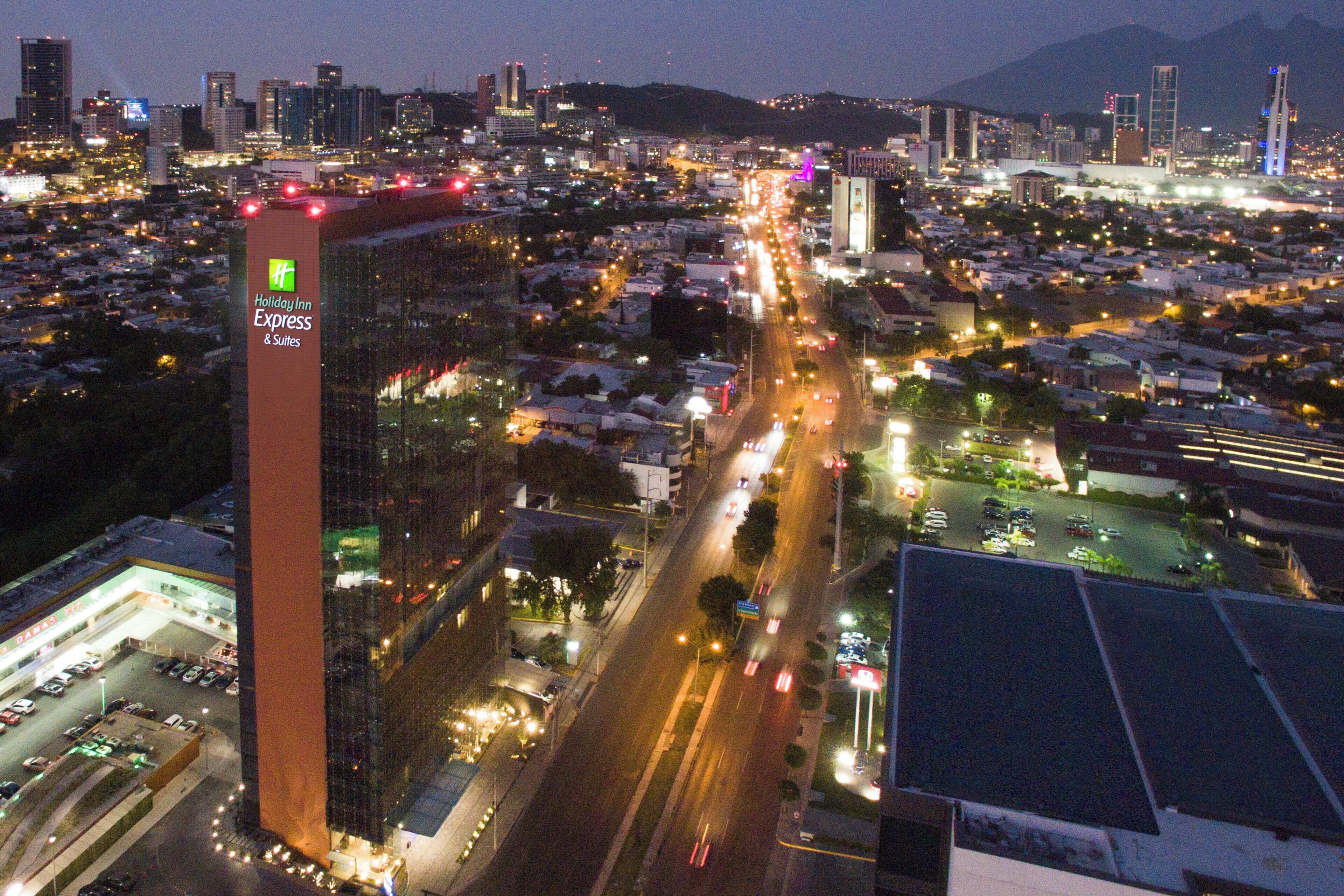 A cityscape at night with a prominent hotel tower labeled 'Holiday Inn Express & Suites' with a parking lot and a road with vehicles.