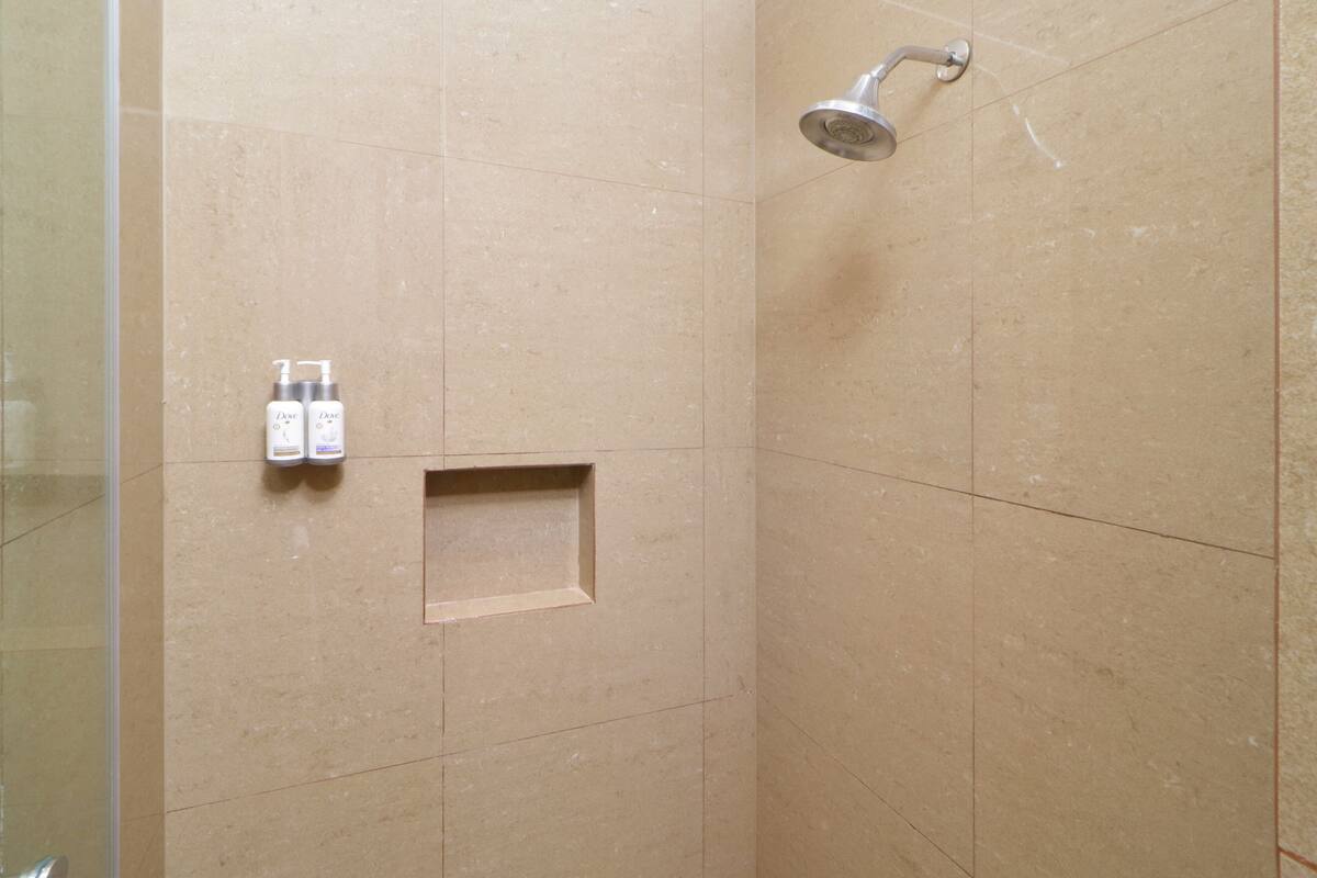 A shower area with beige tiles, a showerhead, and two bottles of Dove body wash on the wall.