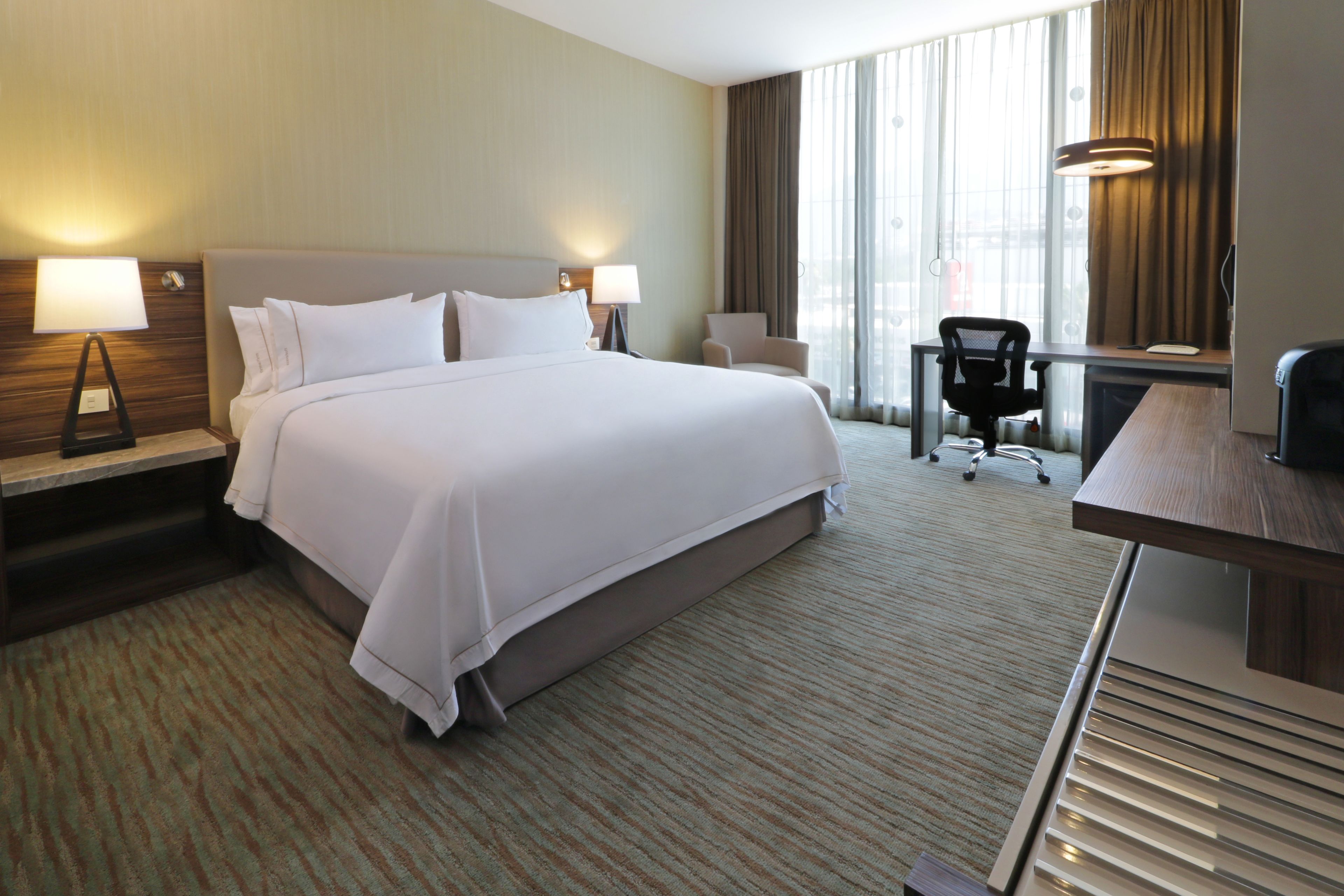 A hotel room with a large bed, two bedside tables with lamps, a desk, a chair, and a large window with curtains.