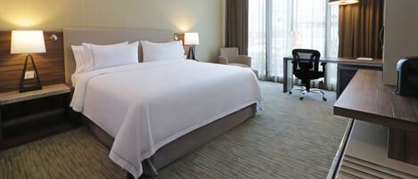 A hotel room with a large bed, two bedside tables with lamps, a desk, a chair, and a large window with curtains.
