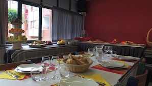 Una mesa de comedor dispuesta con platos, vasos y pan, con una barra de frutas y verduras al fondo.