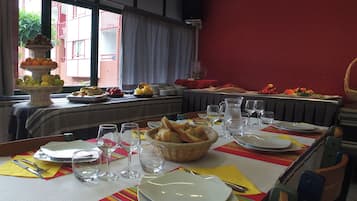 Una mesa de comedor dispuesta con platos, vasos y pan, con una barra de frutas y verduras al fondo.