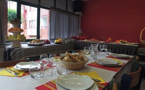 Mesa de jantar posta com pratos, copos e pão, com um buffet de frutas e legumes ao fundo.