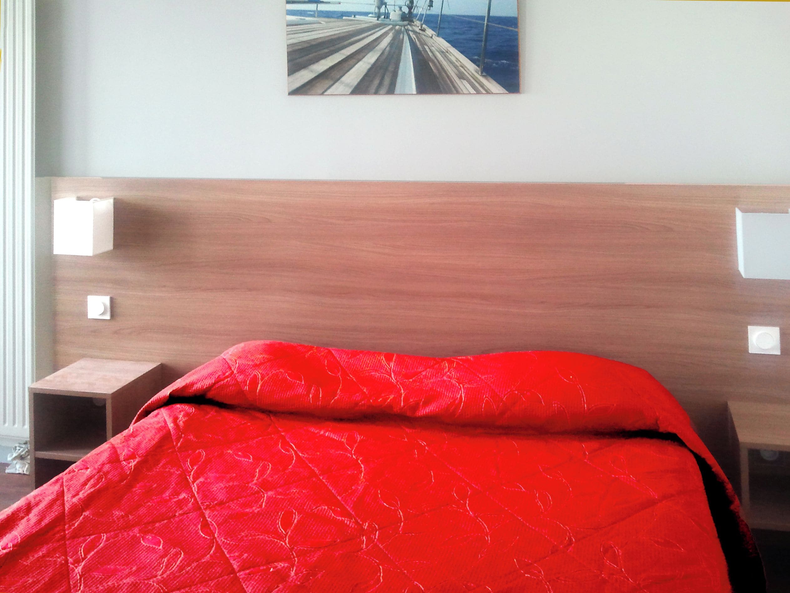 Una habitación de hotel con cabecera de madera, colcha roja y un cuadro en la pared.